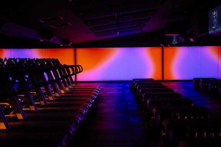 A spacious fitness studio with a row of treadmills and purple and red lit walls.